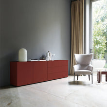 Laden Sie das Bild in den Galerie-Viewer, Sideboard in kräftigem English Red in einer modernen Wohnumgebung mit grauer Wand, hellem Boden und bequemen Sessel am Fenster.