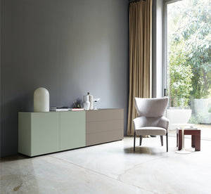 Moderne Sideboardkombination mit Piure Nex Pur Box Türmodul in Salbeigrün links und Schubkastenelement in warmem Taupegrau rechts, in heller Wohnumgebung mit grauer Wand und Design-Sessel.