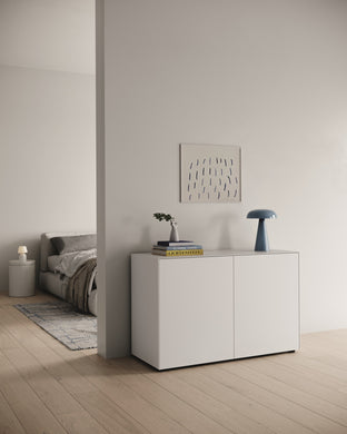 Kompakte Nex Pur Box mit zwei Türen in Weiß, freistehend vor einer Wand zwischen Schlafzimmer und Flur, mit Deko und Designlampe, auf hellem Holzfußboden – moderner, luftiger Look.