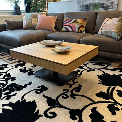 Design-Couchtisch Ronald Schmitt Janus K472 Lignum mit massiver Schublade in Wildeiche natur und vertikaler Rillenstruktur – geschlossen im Wohnambiente mit Ecksofa und Teppich.