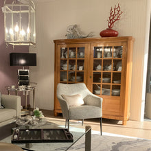 Laden Sie das Bild in den Galerie-Viewer, Form Exclusiv Camargue – Landhausvitrine in Kirschbaum massiv, 155×162×46 cm, Ambienteansicht mit Sessel
