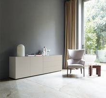 Laden Sie das Bild in den Galerie-Viewer, Wohnsituation mit Nex Pur Box Sideboard in komplettem Seidengrau: links Türmodul 1536778, rechts Schubkastenelement 1536776, vor grauer Wand mit großer Fensterfläche und Designstuhl.
