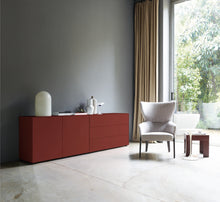 Laden Sie das Bild in den Galerie-Viewer, Sideboard in kräftigem English Red in einer modernen Wohnumgebung mit grauer Wand, hellem Boden und bequemen Sessel am Fenster.
