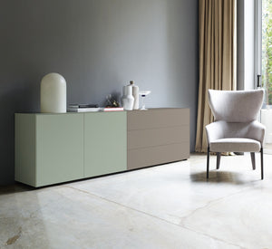 Moderne Sideboardkombination mit Piure Nex Pur Box Türmodul in Salbeigrün links und Schubkastenelement in warmem Taupegrau rechts, in heller Wohnumgebung mit grauer Wand und Design-Sessel.
