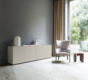 Seidengraue Sideboardkombination in einer stilvollen Einrichtung mit grauer Wand, Vorhängen und Sessel neben großem Fenster, mit minimalistischer Dekoration.